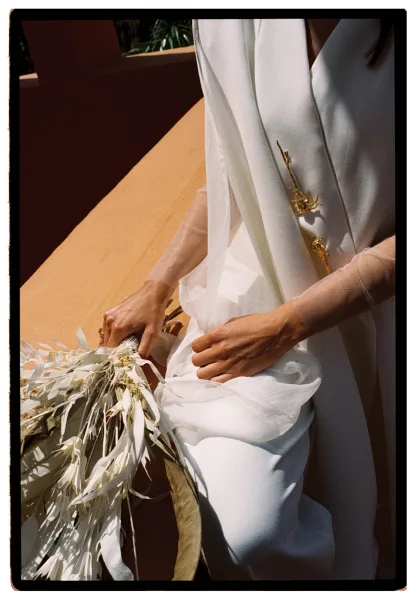 Bridal portrait of a bride holding bouquet with ribbon, wearing a veil and gold hair accessory, seated on a sunlit stone bench by greenery