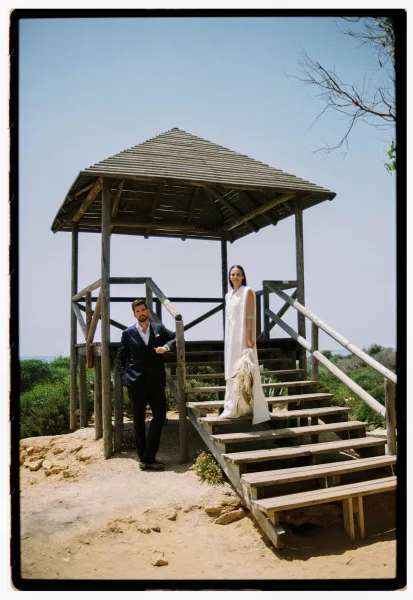 Couple portrait of bride in a sleeveless gown holding a dried flower bouquet beside groom in a black suit on wooden gazebo stairs outdoors