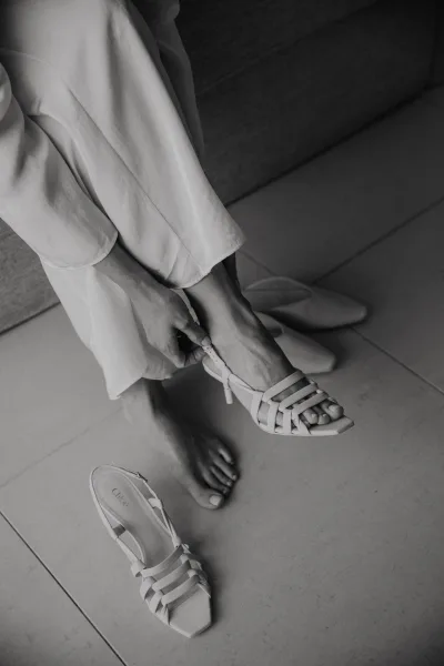 Bridal shoes, white wedding heels with strappy accents beside a bridal robe on a tiled floor near an upholstered chair