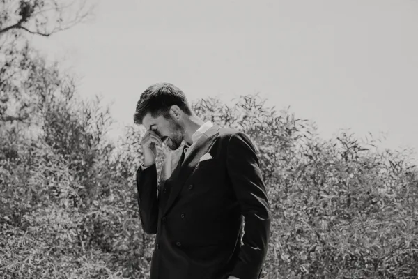 Groom portrait in a black and white wedding portrait, wiping tears in a tuxedo with pocket square against trees and sky backdrop