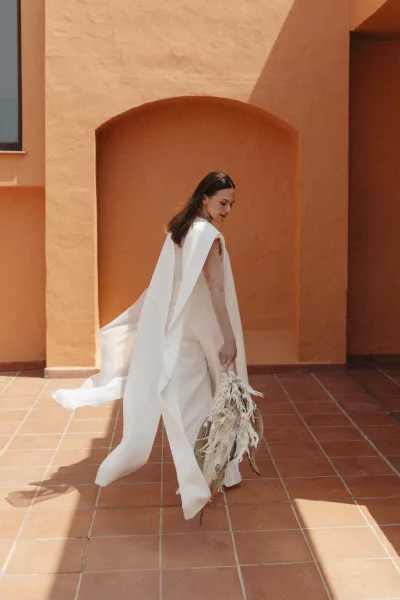 Bridal portrait of a bride in a wedding dress with cape veil holding a dried flower bouquet beneath a stucco archway courtyard