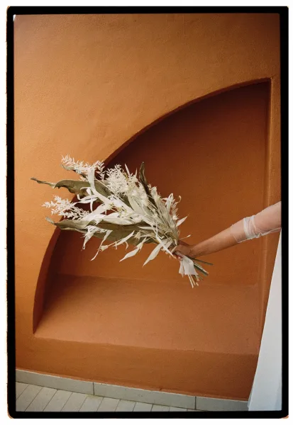 Bridal bouquet of white dried florals and greenery with ribbon wrap, held in sheer gloves against a terracotta stucco niche backdrop