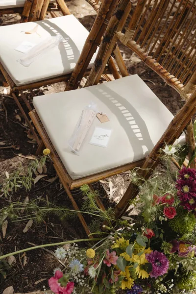 Ceremony chair details with rattan chairs and cushions, wedding programs and favor bags with gift tags beside colorful aisle florals outdoors