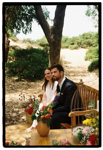 Couple portrait of bride and groom sitting on a wooden bench with colorful florals accent in an outdoor garden setting