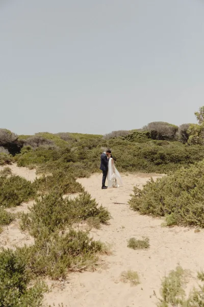 Wedding couple portrait of bride and groom kissing on sand dunes, her veil blowing as she holds a bouquet under open sky