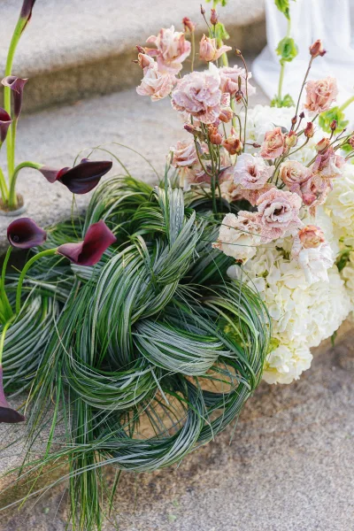 Wedding bouquet with burgundy calla lilies, blush lisianthus, white hydrangea, and cascading greenery on stone steps by a white drape