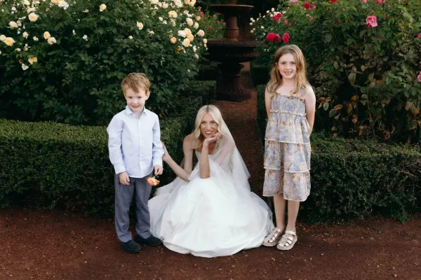 Bride with children smiling in a garden bridal portrait, her classic veil catching light beside blooming rose bushes near a fountain.