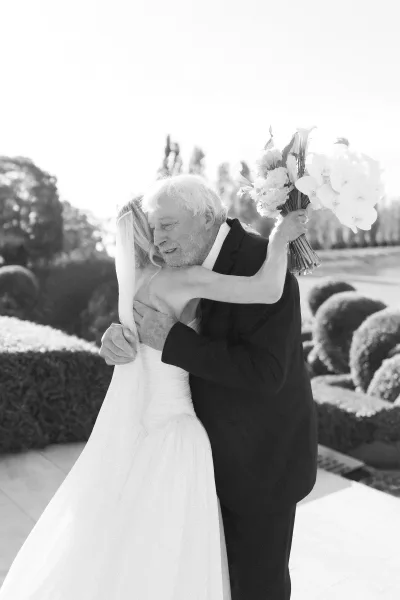 Father daughter moment as a bride hugs her father, clutching an orchid bouquet on a garden terrace framed by hedges, topiary, and trees.