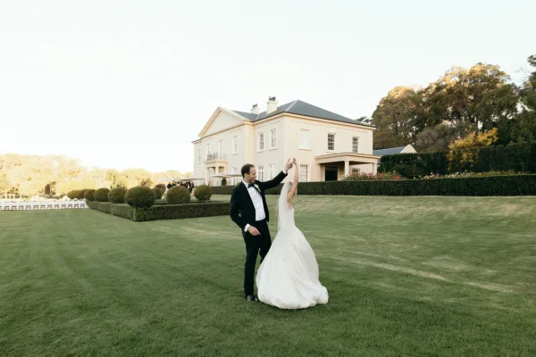 Couple portrait with bride twirling in gown, her veil sweeping as the groom stands beside her on a manicured lawn by an estate house.
