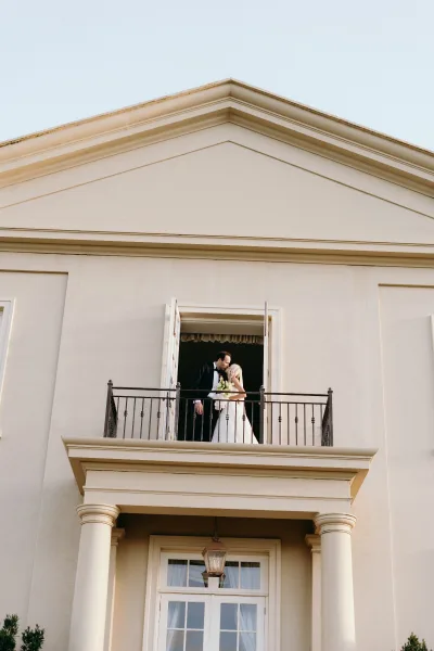 Couple portrait of a bride and groom on a small balcony, the bride holding a bouquet, framed by classic columns against a stately building facade.