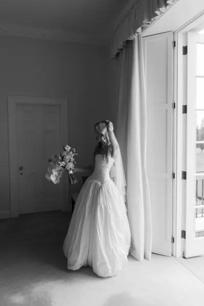 Bridal portrait of a bride by window, wearing a strapless ball gown and holding a classic bouquet in soft window light.