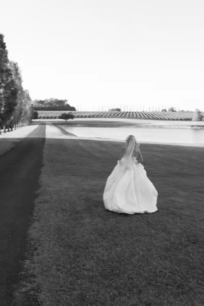 Bridal portrait of a bride walking away, her long veil trailing as she crosses a grass lawn toward a lakeside path framed by vineyard rows.