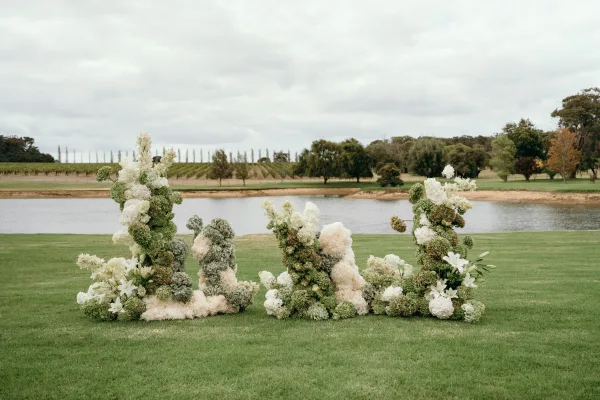 Ceremony backdrop featuring grounded ceremony florals of white hydrangeas on a grass lawn by a lake, with trees and vineyard rows beyond.