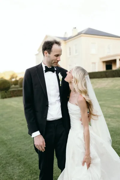 Couple portrait of bride and groom smiling, the groom in a classic tux with a calla lily boutonniere, on the mansion lawn under open sky.