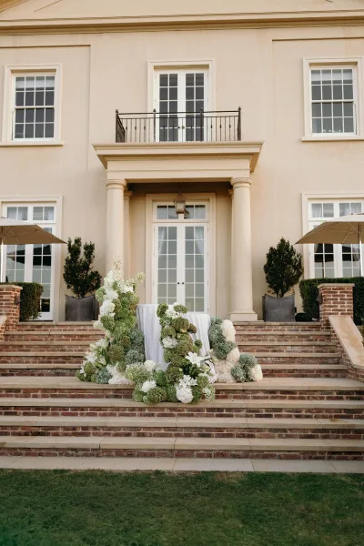 Outdoor ceremony decor with staircase wedding florals framing brick steps, featuring hydrangeas, set before a mansion facade and French doors.