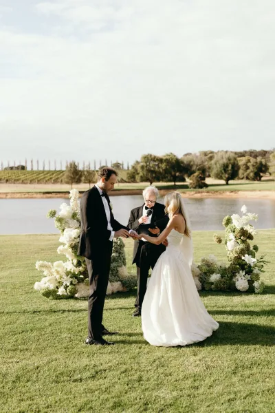 Ring exchange during an outdoor ceremony moment as the groom places the ring on the bride, framed by green and white florals at a lakeside vineyard.