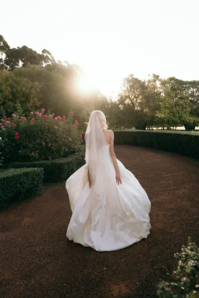Bridal portrait of a bride walking the garden path under sunlit trees, veil trailing over her gown between tall hedges