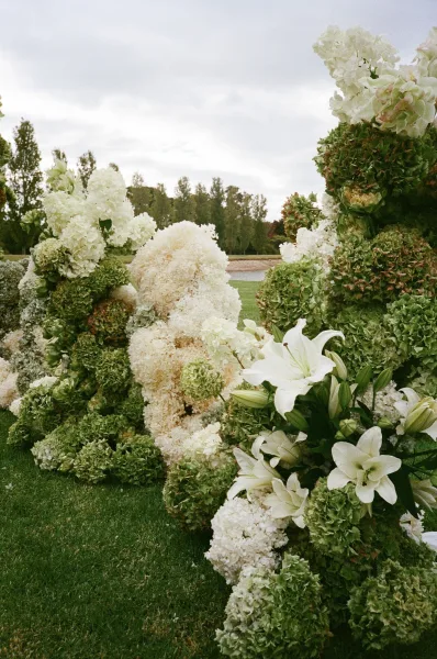 Ceremony flowers create an outdoor ceremony floral installation featuring lush hydrangeas on a grass lawn, with trees behind and a soft cloudy sky.