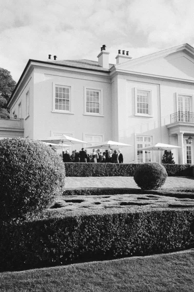 Outdoor cocktail hour at a garden cocktail reception, guests mingling under umbrellas on a manor house terrace with hedges and topiary.