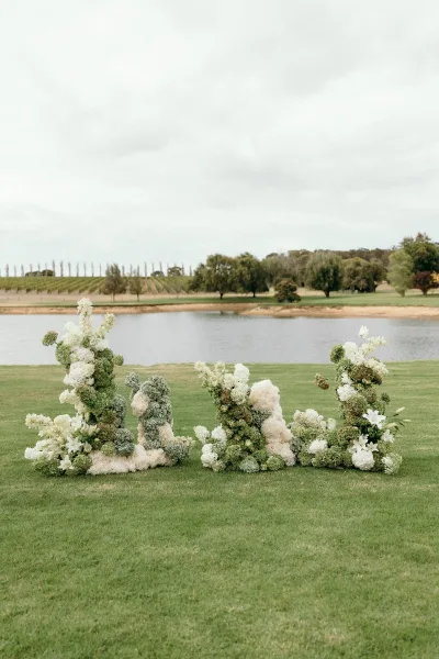 Wedding altar flowers frame the outdoor ceremony decor, low clustered blooms with lilies set on a grass lawn by a lakeside vineyard under clouds.