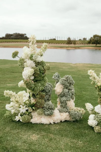 Ceremony flowers shape outdoor ceremony decor, hydrangeas gathered on a grass lawn by a lakeside vineyard beneath a cloudy sky.
