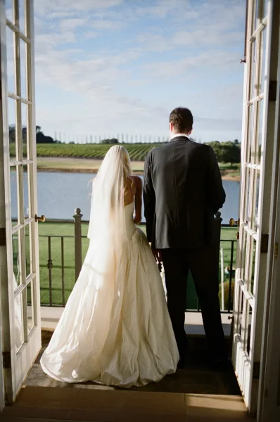Couple portrait of bride and groom on balcony, her long veil catching light as they overlook a serene lake and vineyard under open sky.