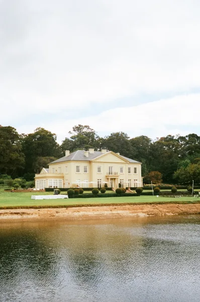 Estate wedding venue on a mansion wedding venue’s lakeside lawn, warm string lights glow as trees frame the scene under a cloudy sky.