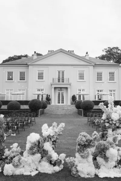 Ceremony setup showcasing elegant outdoor ceremony decor on a mansion lawn, with topiary shrubs lining stone steps beneath an open sky.