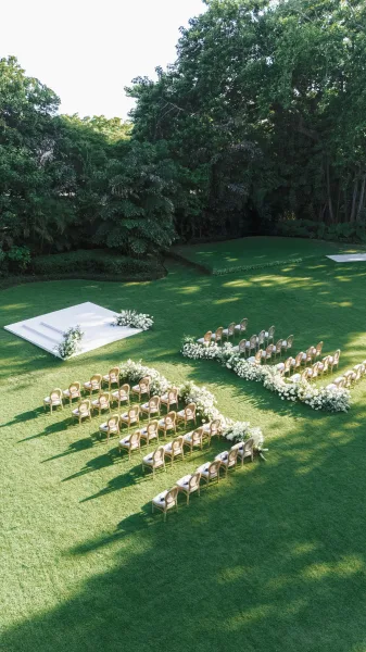 Ceremony setup with a white aisle runner leading to a white stage platform, low white florals, and curved chairs on a sunlit lawn