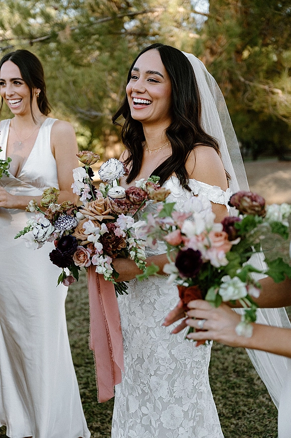 Bride portrait of a smiling bride holding bouquet in an off-the-shoulder lace gown with veil, with trees and greenery behind her