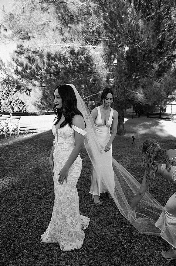 Bride getting ready as bridesmaids help adjust her bridal veil over a lace off-the-shoulder gown on a sunny lawn with trees