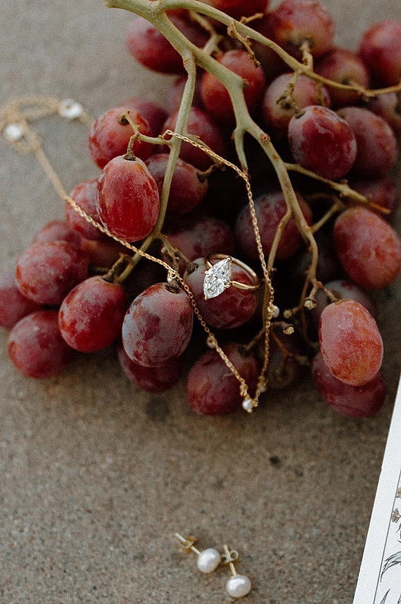 Engagement ring and pear shaped engagement ring set with pearl earrings, gold necklace, grapes, and paper card on a stone surface