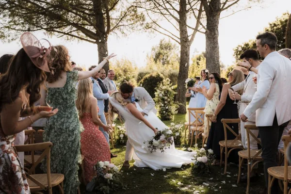 Wedding kiss moment during the ceremony recessional as newlyweds dip-kiss in a sunlit garden aisle, guests cheering beside floral chairs