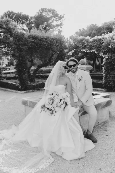 Couple portrait, a black and white wedding portrait of bride and groom on a stone bench in a garden, holding a bouquet with veil flowing