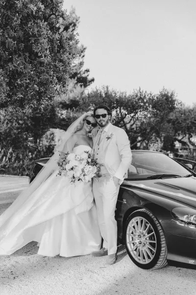 Couple portrait of bride and groom sunglasses, leaning on a car on a gravel driveway, bride in veil holding bouquet, groom in white suit
