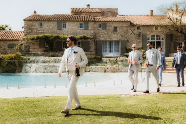Groomsmen portrait of the wedding party walking in sunglasses, suits and boutonnieres beside a stone villa pool under blue sky
