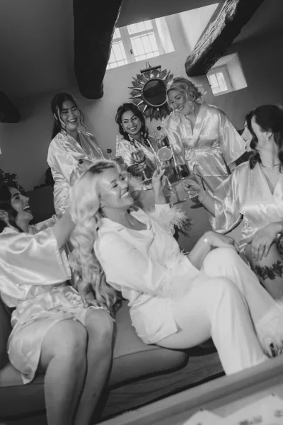 Bridesmaids getting ready share a bridal party toast in satin robes, laughing on a couch with champagne flutes in a bright suite