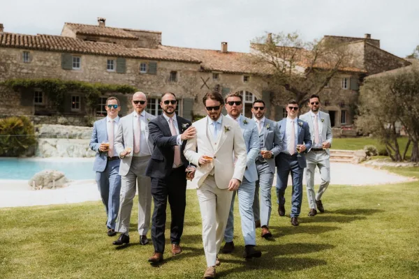 Groomsmen portrait of groom and friends in mix-and-match suits with boutonnieres, sunglasses, and whiskey glasses by a stone villa pool