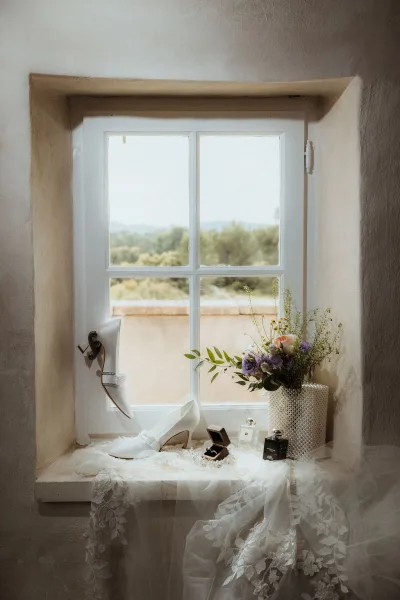 Bridal details wedding detail flat lay on a window ledge with white heels, lace veil, bouquet with greenery, rings, pearl bag, and perfume bottle