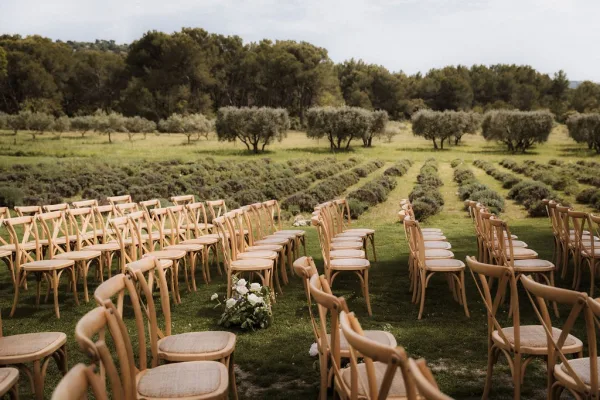 Outdoor ceremony setup with wedding ceremony seating of wood chairs along a curved aisle, with white florals on a lawn by lavender fields and olive trees