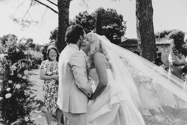 Wedding couple portrait in a black and white wedding photo, bride and groom laughing in a garden as her veil lifts near guests and stone building