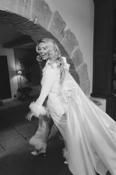 Bridal portrait in black and white of a laughing bride in a white jumpsuit with feather-trim cape, standing in a stone archway hallway