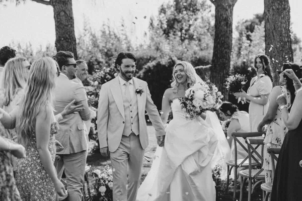 Ceremony recessional as newlyweds walking down aisle holding hands, bride with bouquet and veil, groom in suit amid petal confetti in a garden setting