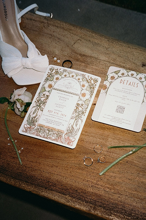 Invitation suite with floral wedding invitations, RSVP QR code card, rings and pearl earrings beside a white bridal shoe on a wood table