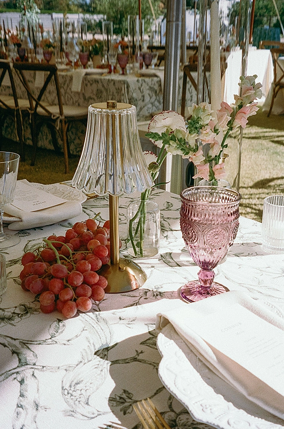 Reception tablescape wedding table setting with patterned tablecloth, pleated glass lamp, pink goblet, florals and grapes in a sunlit tent