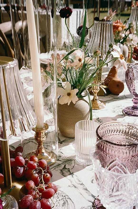 Reception tablescape with wedding table centerpiece of taper candles in brass candlesticks, floral vase, grapes and pears on patterned linen tablecloth