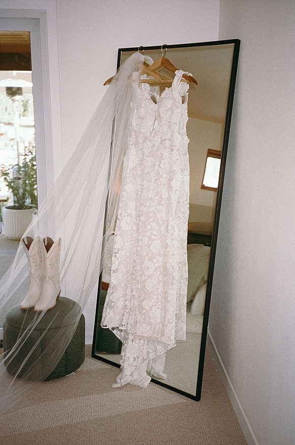 Wedding dress with lace wedding dress details hanging on wooden hanger, long veil beside cowboy boots in a bright bedroom by a window