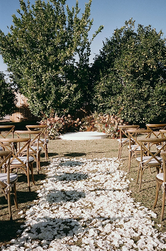 Outdoor ceremony setup with wood crossback chairs and garden wedding ceremony aisle lined in rose petals, leading to a circular platform on a lawn under trees