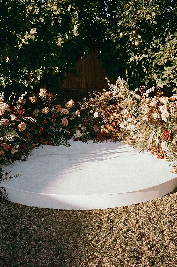 Wedding ceremony backdrop with roses and greenery garland on a white platform stage, set against a leafy hedge and wood gate in sunlight