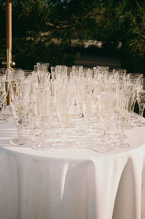 Champagne glassware and wedding champagne flutes displayed on a white tablecloth with evergreen branches in the background outdoors